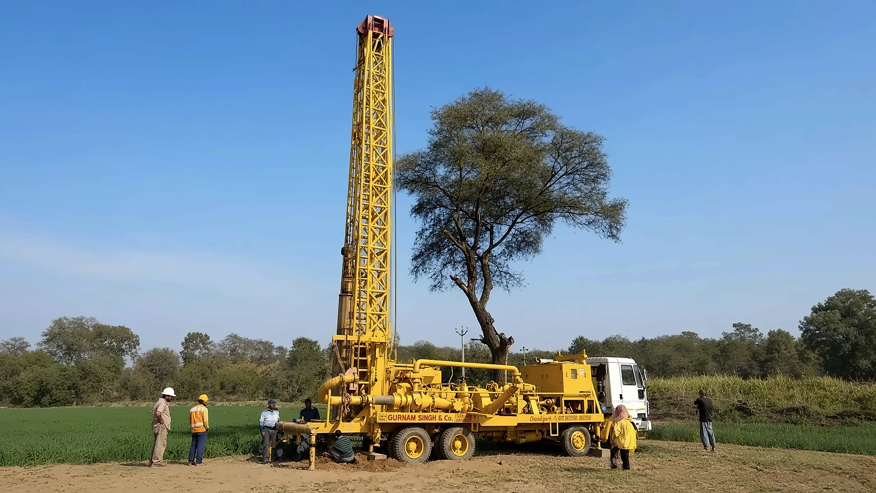 A Gurnam Singh and Company rig at work in a field, mast raised, crew at the drill table