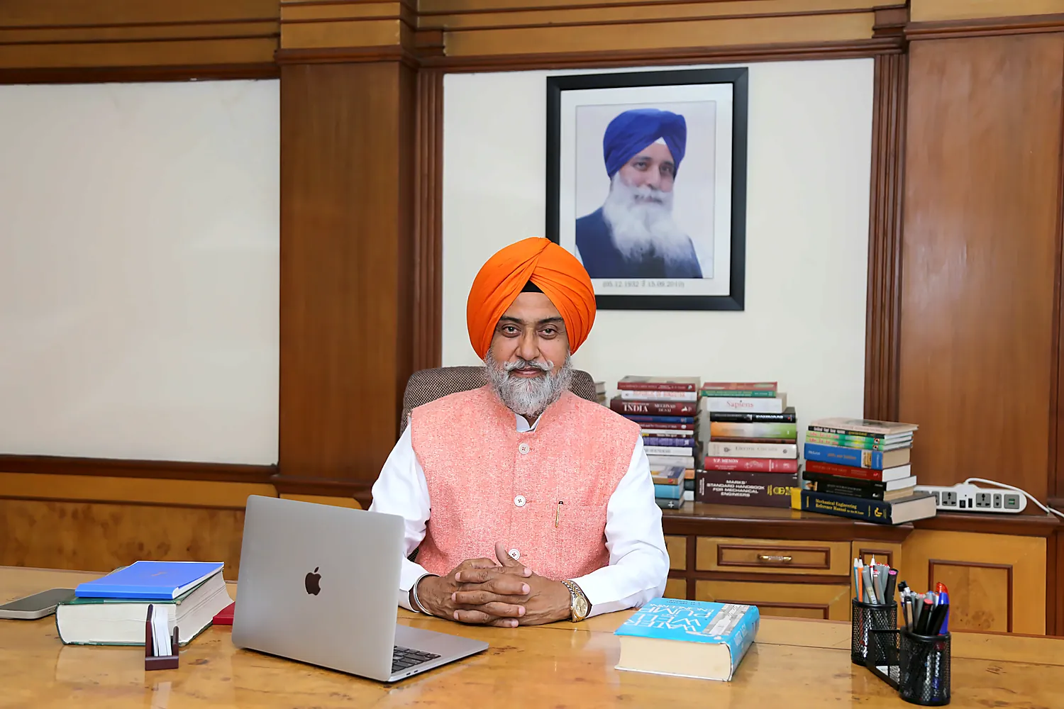Sardar Gurdarshan Singh Saini at his desk, beneath a portrait of the founder